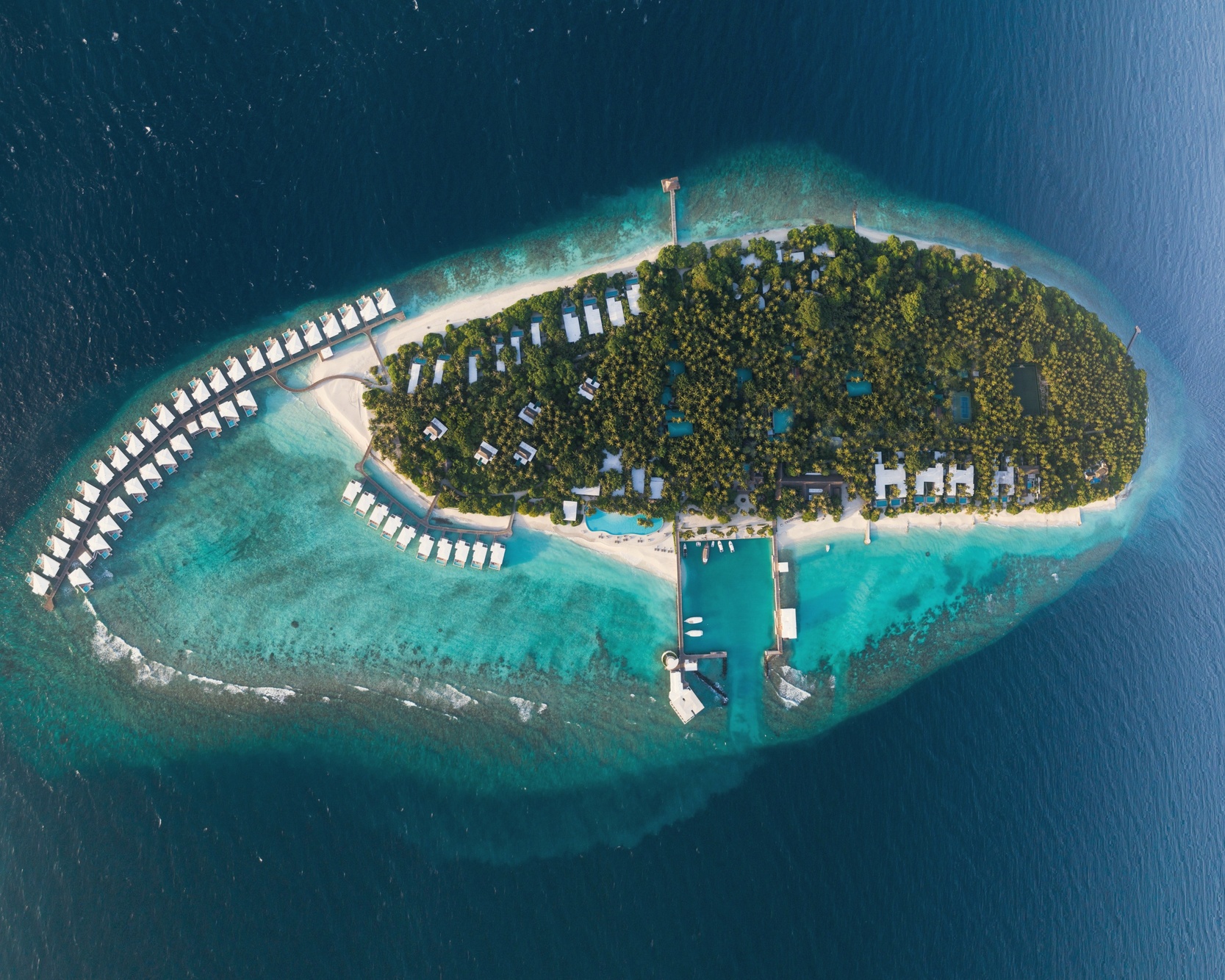 Finolhas Island from above, the overwater villa jetties extending into the Baa Atoll lagoon, the UNESCO reserve waters visible beyond.