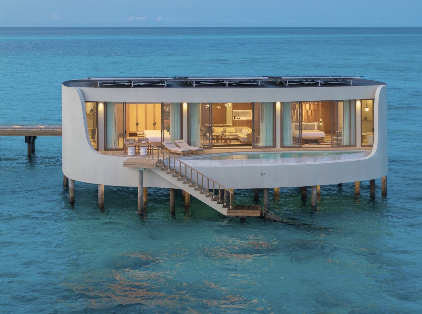 The interior of a sunset-facing overwater pool villa. Warm light across the water at the end of the day, with the pool deck oriented west.