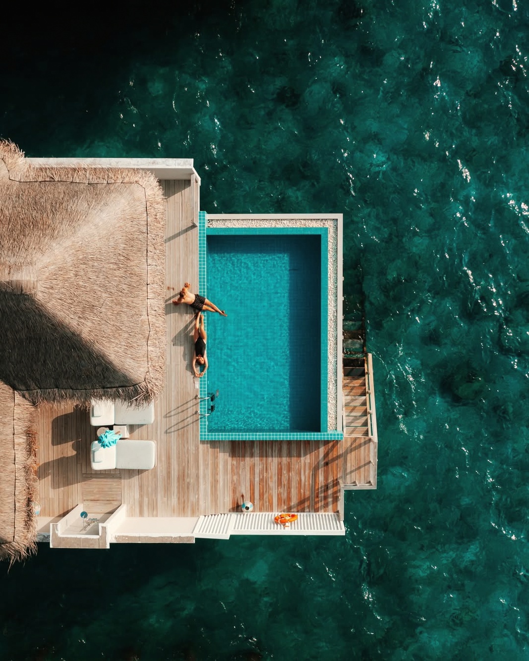 An overwater villa from the deck, pool at the edge, the lagoon below. The water in Baa Atoll is clear at this depth.