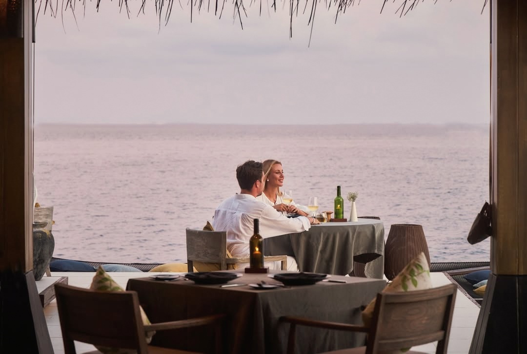 The main restaurant at water level. Tables set for dinner with the lagoon visible on three sides as the light drops.