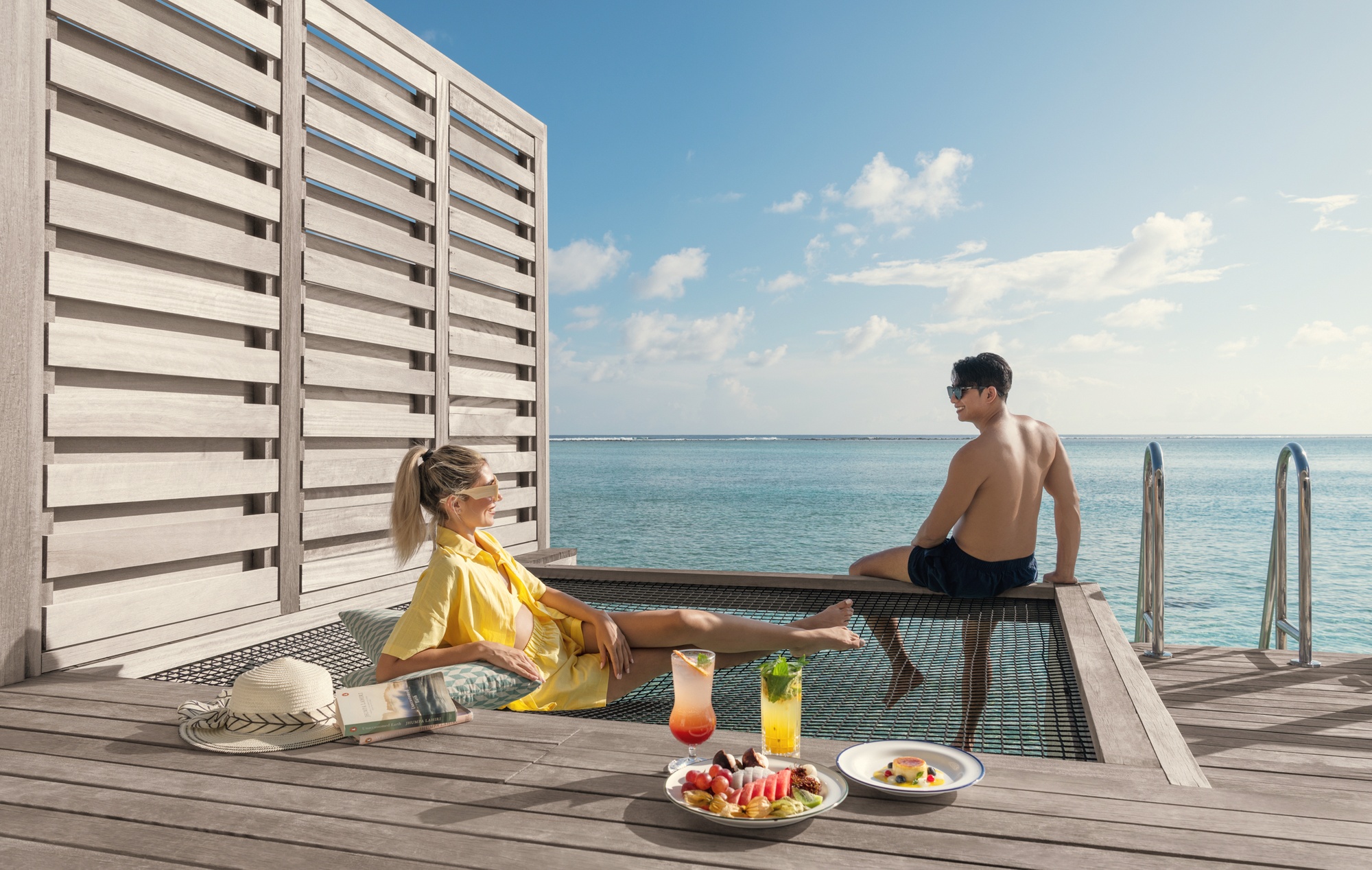 An overwater villa extending over the lagoon. The water below is clear and shallow, with reef visible from the deck.