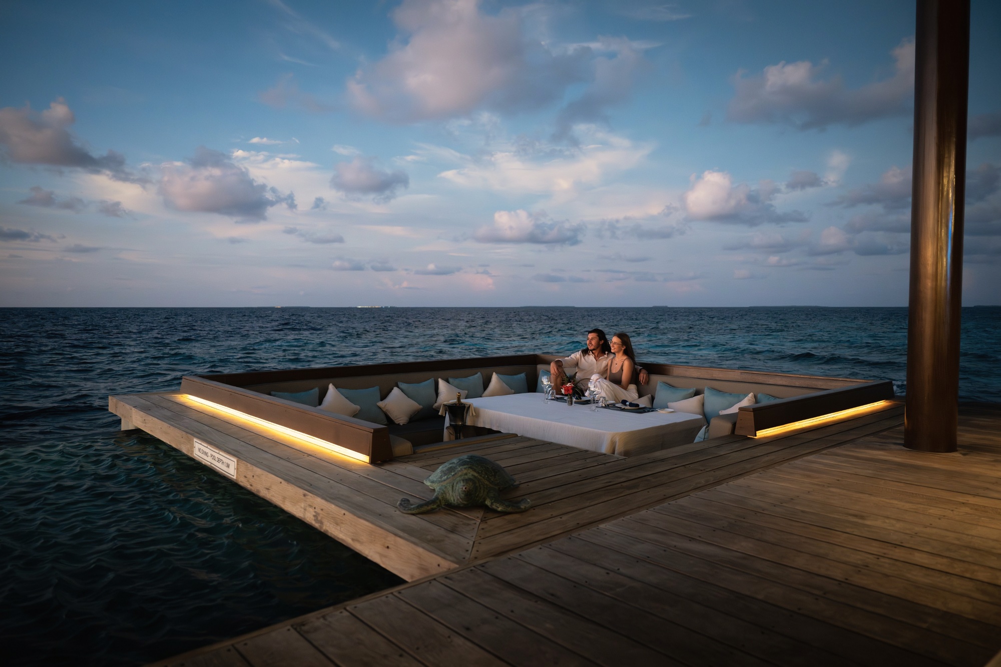 An overwater villa from the deck. The lagoon edge extends directly from the villa floor, with the atoll's characteristic deep-water clarity visible below.