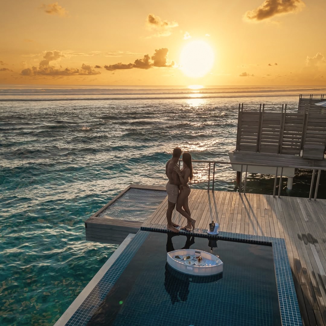 An overwater villa deck at water level. The lagoon floor is visible from the deck, and the South Ari reef system begins within swimming distance.