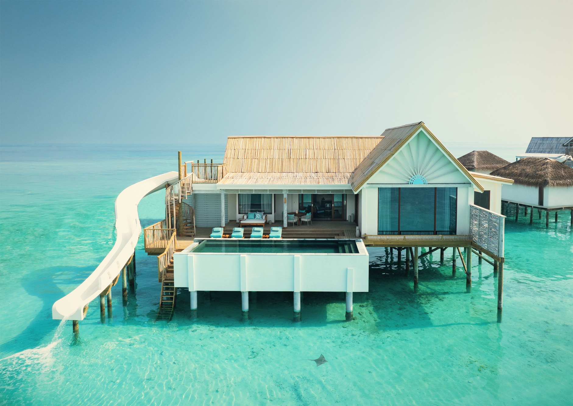 An overwater pool villa deck at dusk. The water below is clear and shallow, with direct lagoon entry from the villa.