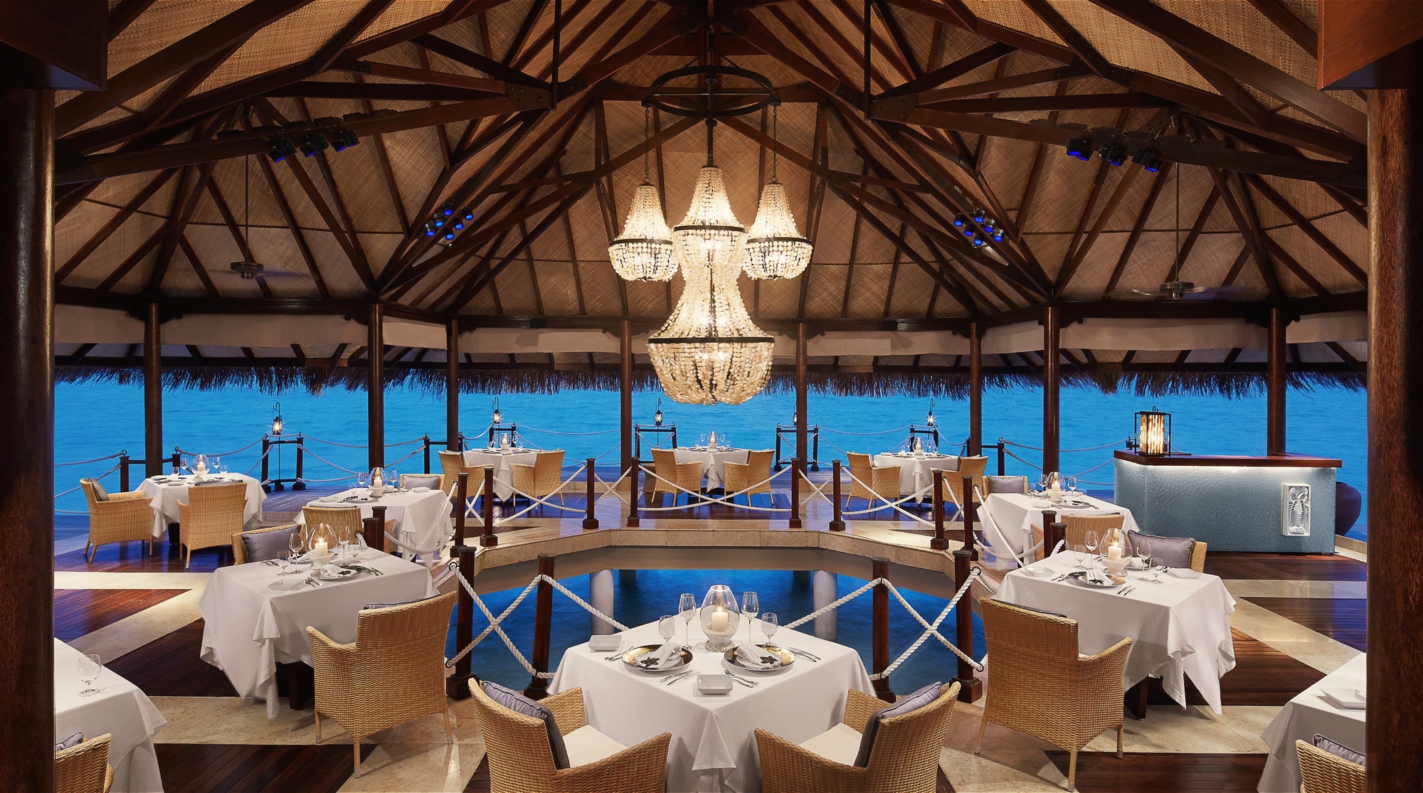 The resort's main restaurant at dinner. Tables set for the evening service, the lagoon visible beyond the open-sided dining room.